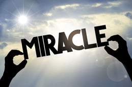 Two hands holding up the letters spelling the word Miracle with the blue sky behind them.